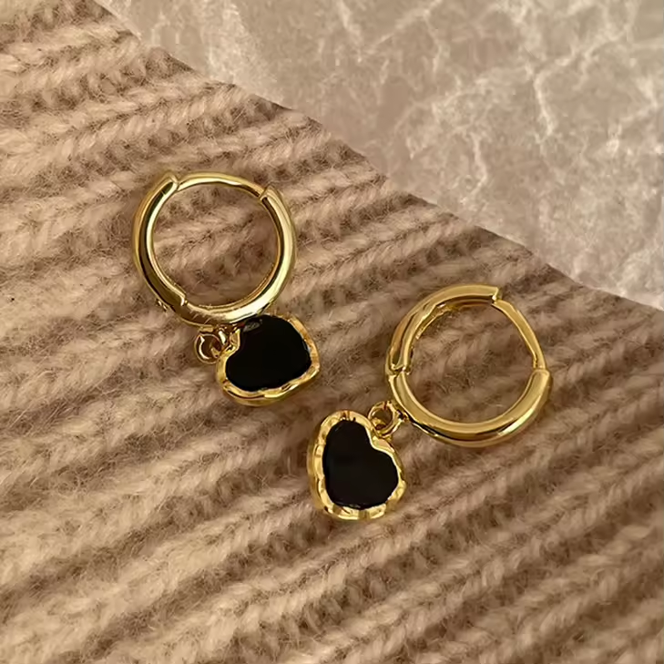 Elegante Huggies-Ohrringe aus 925 Sterling Silber mit schwarzem Herzanhänger, vergoldet mit 18K, auf weichem Stoffhintergrund.