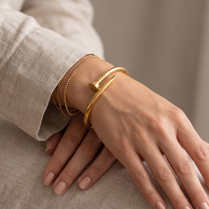 Ein eleganter, goldener Nagel-Armreif der Marke BOGUE, getragen am Handgelenk einer Frau in Kombination mit feinen Perlenarmbändern auf beigem Leinenstoff.
